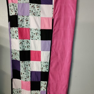May include: A patchwork quilt with a black, white, pink, purple, and green color scheme. The quilt is made of squares with different patterns. The quilt is hanging on a wall with a solid pink fleece blanket hanging next to it.
