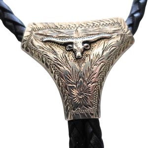 May include: A bolo tie featuring a silver-toned metal slide with an engraved longhorn steer head and floral patterns. The slide is attached to a black braided leather cord that extends upwards and downwards from the central decorative piece.