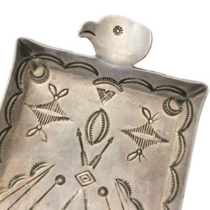 May include: A silver-toned metal decorative tray with an eagle head at the top. The tray features etched geometric designs, including arrows, diamonds, and starburst patterns. The edges are scalloped.