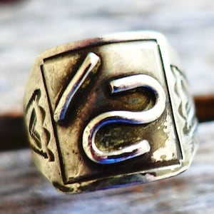 Puede incluir: Un anillo de plata con un diseño cuadrado que presenta el número "15" en una fuente estilizada.