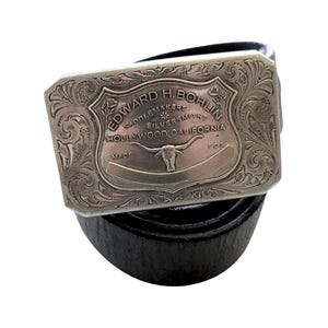 Puede incluir: Un cintur&oacute;n de cuero negro con una hebilla rectangular plateada. La hebilla presenta un dise&ntilde;o grabado con el texto "EDWARD H. BOHLIN SADDLEMAKERS SILVERSMITHS HOLLYWOOD, CALIFORNIA" y una imagen de un toro.