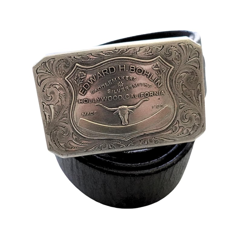 Puede incluir: Un cintur&oacute;n de cuero negro con una hebilla rectangular plateada. La hebilla presenta un dise&ntilde;o grabado con el texto "EDWARD H. BOHLIN SADDLEMAKERS SILVERSMITHS HOLLYWOOD, CALIFORNIA" y una imagen de un toro.