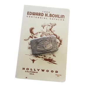 Puede incluir: Una hebilla de cintur&oacute;n plateada con un dise&ntilde;o grabado con un longhorn, apoyada en un cat&aacute;logo. El cat&aacute;logo es de color crema y tiene el texto "EDWARD H. BOHLIN CENTENNIAL CATALOG" y "HOLLYWOOD CALIFORNIA U.S.A."