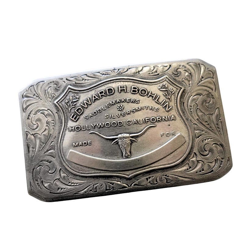 Puede incluir: Hebilla de cintur&oacute;n rectangular plateada con adornos de volutas y el texto "EDWARD H. BOHLIN SADDLEMAKERS SILVERSMITHS HOLLYWOOD, CALIFORNIA". Un toro de cuernos largos est&aacute; centrado en la hebilla.