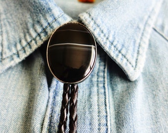 Puede incluir: Un bolo tie de ágata negro y blanco con un cordón de cuero trenzado. El bolo tie está sobre una camisa vaquera.