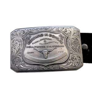 Puede incluir: Hebilla de cintur&oacute;n plateada con intrincados adornos y el texto "EDWARD H. BOHLIN SADDLEMAKERS SILVERSMITHS HOLLYWOOD, CALIFORNIA". La hebilla presenta un emblema de toro de cuernos largos.