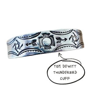 May include: A silver cuff bracelet featuring stamped designs of a central thunderbird, arrows, and geometric symbols. The metal surface is polished with dark oxidised detailing in the engravings. Text on the image reads: TOM DEWITT THUNDERBIRD CUFF.