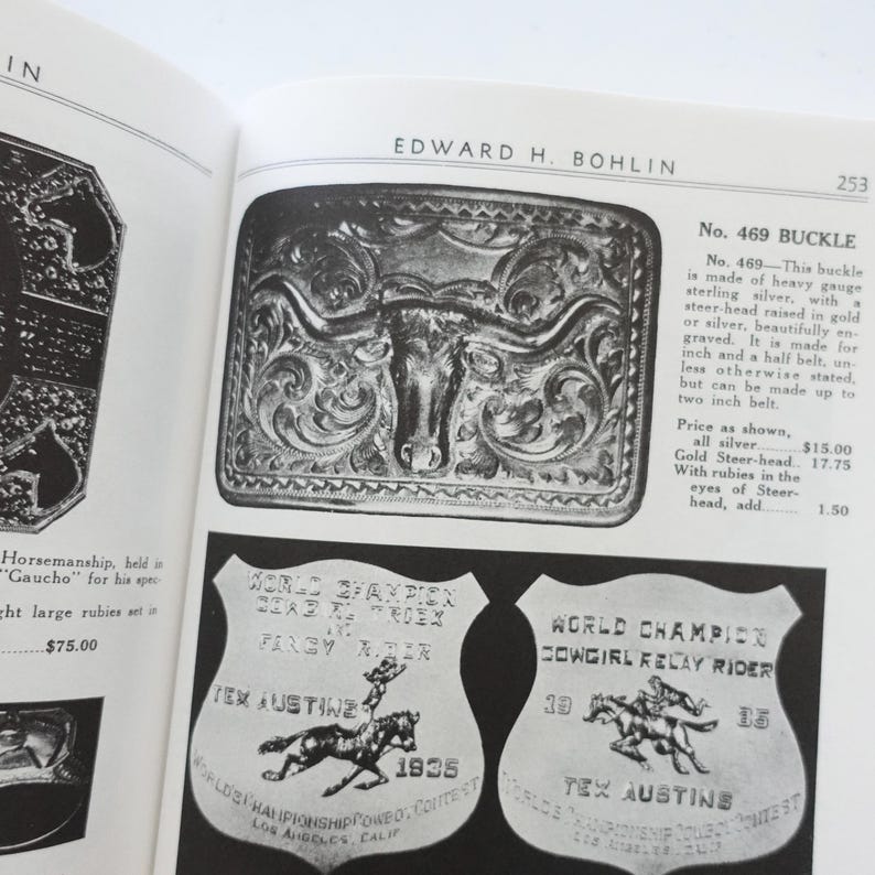 Puede incluir: Una p&aacute;gina de libro abierta con una hebilla de cintur&oacute;n plateada con un dise&ntilde;o de cabeza de toro y dos insignias de campe&oacute;n de rodeo vintage. La hebilla est&aacute; grabada y las insignias tienen texto e ilustraciones de caballos y jinetes.