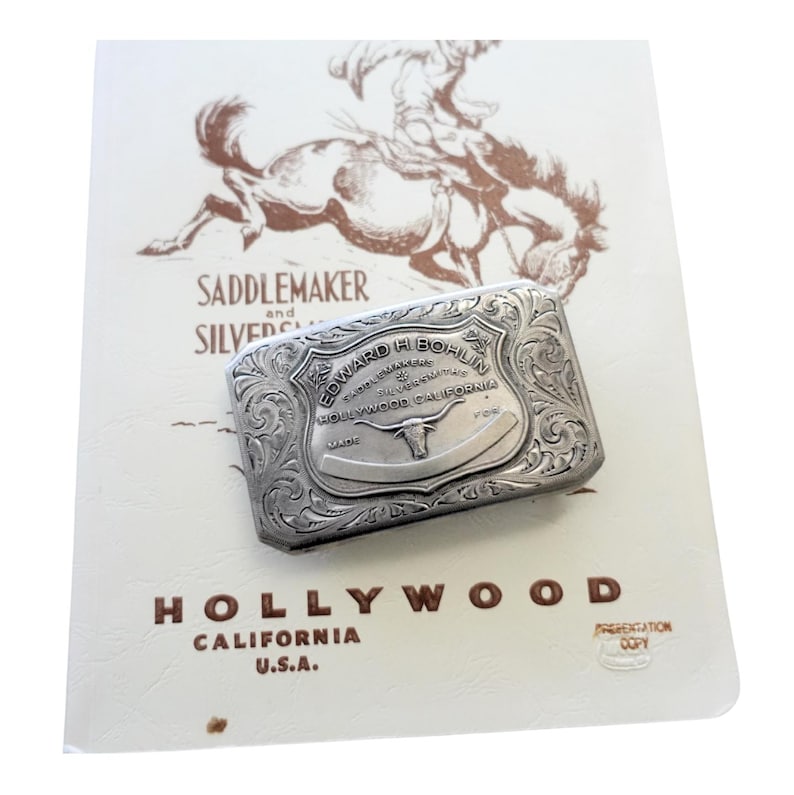 Puede incluir: Un clip de dinero plateado con intrincados dise&ntilde;os florales y de volutas, con el texto "EDWARD H. BOHLIN HOLLYWOOD CALIFORNIA" y un emblema de cuerno largo. El clip descansa sobre un papel color crema con una ilustraci&oacute;n de vaquero y las palabras "SADDLEMAKER and SILVERSMITH".