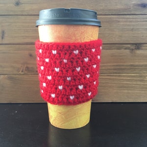 May include: Red crocheted coffee cup cozy with white hearts. The cozy is on a paper coffee cup with a black lid that says "Contenido Caliente".