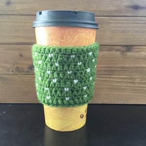 May include: Green crocheted coffee cup cozy with white hearts. The cozy is on a yellow paper coffee cup with a black lid.