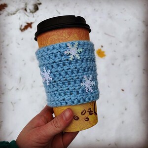 May include: A blue crocheted coffee cup cozy with snowflake embellishments. The cozy is on a brown paper cup with a white and brown design.