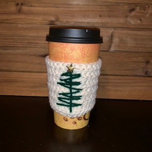 May include: White crocheted coffee cup cozy with a green embroidered Christmas tree and a gold star on a brown paper coffee cup.
