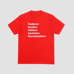 May include: A red t-shirt with the names Fedorov, Kozlov, Fetisov, Larionov, and Konstantinov printed in white. The shirt is made of a soft material and has short sleeves.