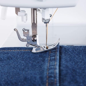 May include: Close-up of a sewing machine needle and foot sewing a seam in blue denim fabric. The needle is threaded with a yellow thread.