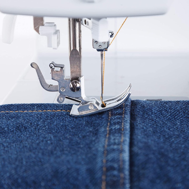 May include: Close-up of a sewing machine needle and foot sewing a seam in blue denim fabric. The needle is threaded with a yellow thread.