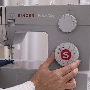 May include: A gray Singer Heavy Duty sewing machine with a white dial for stitch selection and a white dial for stitch length. The machine has a red "S" logo on the front.