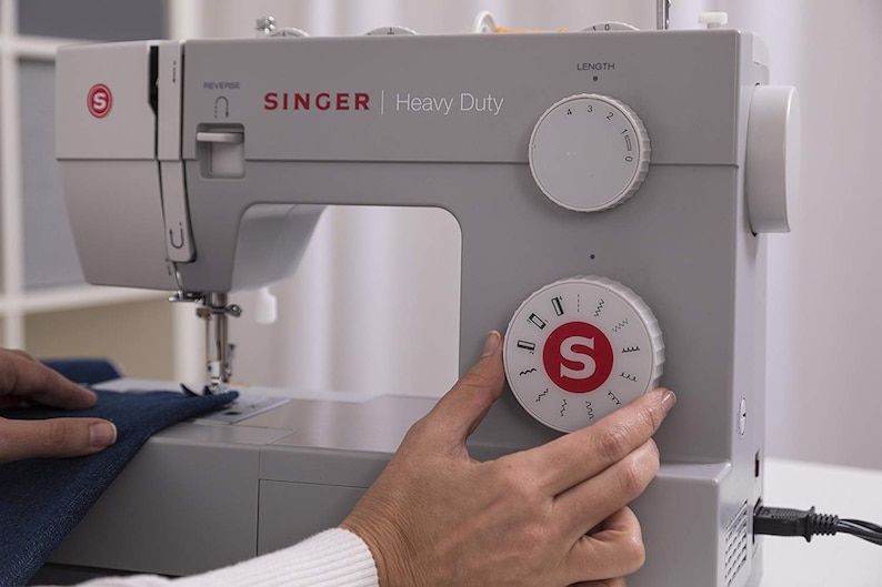 May include: A gray Singer Heavy Duty sewing machine with a white dial for stitch selection and a white dial for stitch length. The machine has a red "S" logo on the front.