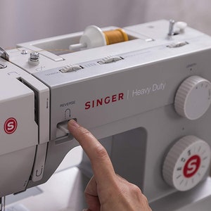 May include: A white Singer Heavy Duty sewing machine with a finger pressing the reverse button. The machine has a spool of thread on the top and a needle in the needle plate. The machine has a stitch selection dial and a tension knob.