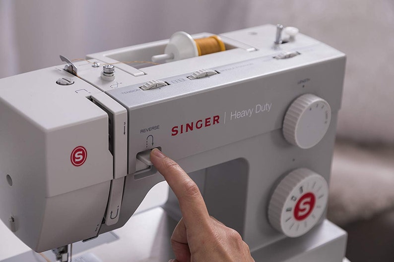 May include: A white Singer Heavy Duty sewing machine with a finger pressing the reverse button. The machine has a spool of thread on the top and a needle in the needle plate. The machine has a stitch selection dial and a tension knob.