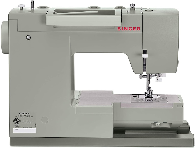 May include: A gray Singer sewing machine with a white foot pedal and a clear plastic cover over the needle. The machine has a white knob on the top right side and a black button on the front. The machine is sitting on a white surface.