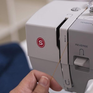 May include: A close-up of a white sewing machine with a brown thread being pulled through the needle. The machine has a red "S" logo and a white knob labeled "Tension".