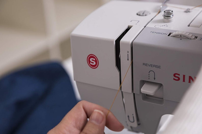 May include: A close-up of a white sewing machine with a brown thread being pulled through the needle. The machine has a red "S" logo and a white knob labeled "Tension".