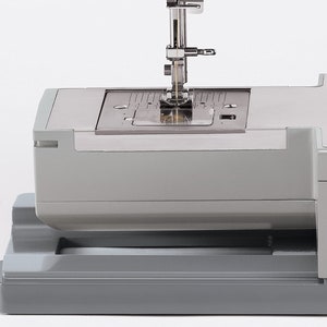 May include: Close-up of a gray sewing machine with a needle and thread in the needle plate. The needle is in the down position and the thread is visible.