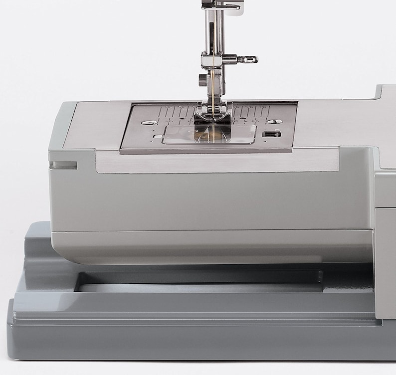 May include: Close-up of a gray sewing machine with a needle and thread in the needle plate. The needle is in the down position and the thread is visible.