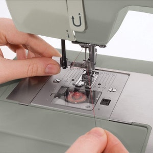 May include: Close-up of a sewing machine with a red thread being threaded through the needle. The needle is in the down position and the thread is being held by a hand.