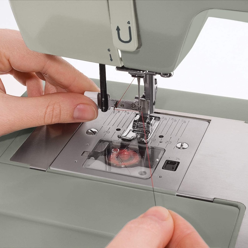 May include: Close-up of a sewing machine with a red thread being threaded through the needle. The needle is in the down position and the thread is being held by a hand.