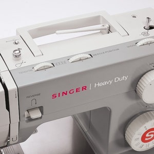 May include: A gray Singer Heavy Duty sewing machine with a white handle and several dials for adjusting stitch length, width, tension, and needle position. The machine has a reverse button and a stitch selection dial.
