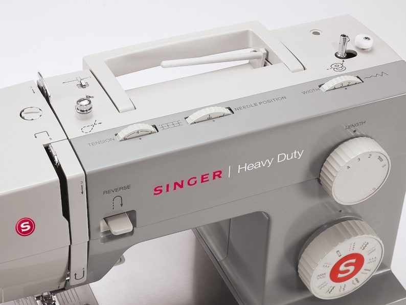May include: A gray Singer Heavy Duty sewing machine with a white handle and several dials for adjusting stitch length, width, tension, and needle position. The machine has a reverse button and a stitch selection dial.