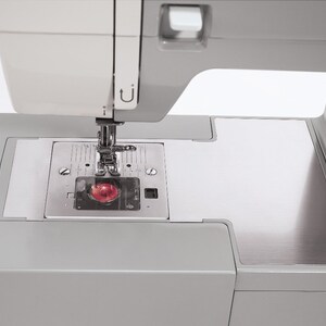 May include: A close-up of a gray sewing machine with a silver needle plate and a red light. The machine has a white top and a gray base.