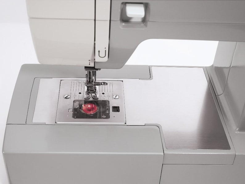 May include: A close-up of a gray sewing machine with a silver needle plate and a red light. The machine has a white top and a gray base.