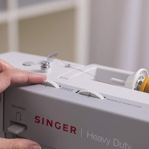 May include: A white Singer Heavy Duty sewing machine with a spool of yellow thread. The machine has a needle position dial, a stitch length dial, and a reverse button.
