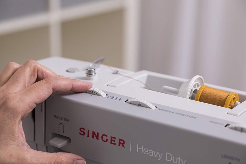 May include: A white Singer Heavy Duty sewing machine with a spool of yellow thread. The machine has a needle position dial, a stitch length dial, and a reverse button.