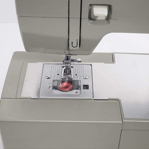 May include: Close-up of a gray sewing machine with a silver needle and a red bobbin. The machine has a clear plastic cover over the bobbin area and a metal plate with markings for stitch length.