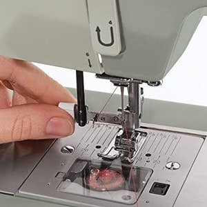 May include: A close-up of a white sewing machine with a needle threaded with red thread. The machine has a clear plastic cover over the needle and bobbin area, and a metal foot pedal.