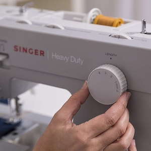 May include: A white Singer Heavy Duty sewing machine with a knob to adjust the stitch length. The knob is labeled with numbers 0 through 4.