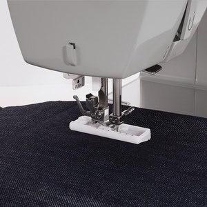 May include: Close-up of a sewing machine needle and foot, with a piece of dark blue denim fabric underneath. The foot is white and has a metal needle guide. The machine has a gray body and a white button labeled "PUSH".