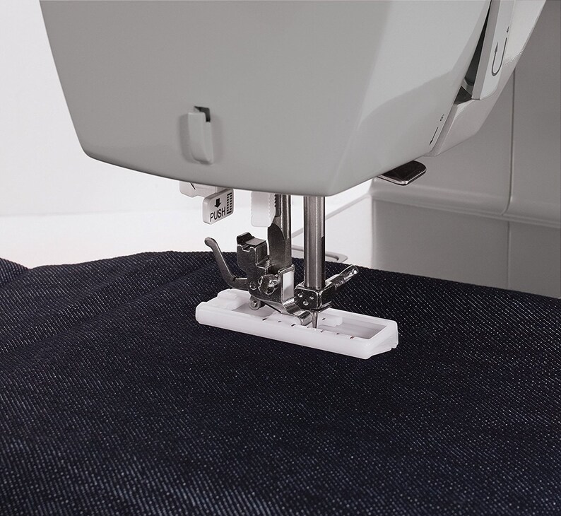 May include: Close-up of a sewing machine needle and foot, with a piece of dark blue denim fabric underneath. The foot is white and has a metal needle guide. The machine has a gray body and a white button labeled "PUSH".