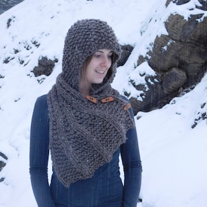 May include: A brown knitted hooded scarf with leather buttons. The scarf is worn by a person in a snowy landscape.