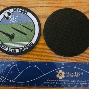 May include: A white, black, and green embroidered patch with the text "SSV-084 USSF ALAN SHEPARD" and a depiction of a spaceship and missiles. The patch is attached to a black velcro backing.