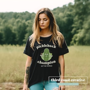May include: Black t-shirt featuring a green cartoon pickle holding a shot glass and a pickleback. The text on the shirt reads "pickleback champion of the world".