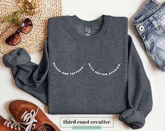 Könnte beinhalten: Ein dunkelgrauer Pullover mit dem Aufdruck "Scars are tattoos... with better stories." in Weiß auf der Vorderseite.
