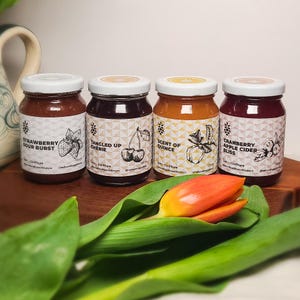 May include: Four jars of fruit preserves with white lids and labels. The labels read "Strawberry Sour Burst", "Tangled Up Cherie", "Scent of Quince", and "Cranberry Apple Cider Bliss". A red and yellow tulip rests in front of the jars.