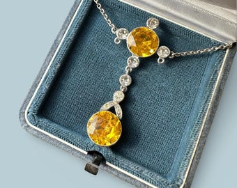 May include: A silver necklace with a pendant featuring a large, round, yellow gemstone at the bottom and a similar gemstone at the top. The necklace is set in a blue velvet jewelry box.