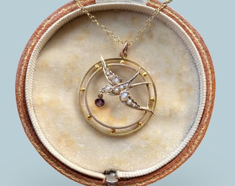 May include: A gold necklace with a pendant featuring a pearl-encrusted swallow in flight, holding a purple gemstone. The pendant is set in a circular frame with a gold border.