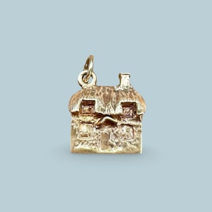 May include: A gold-coloured metal charm shaped like a small cottage with a textured roof, two windows, and a central door. The charm features a small circular loop at the top for attachment, with a detailed, rustic exterior finish throughout.